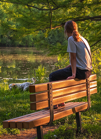 Person sitting on a bench in front of a pond looking at her dog.