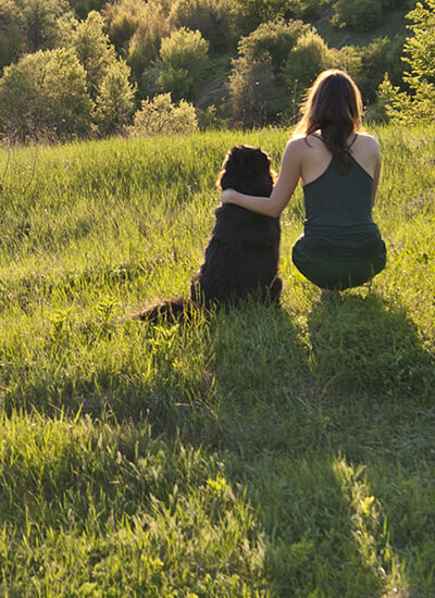 Person and a dog sitting on an open field looking out into an open field.